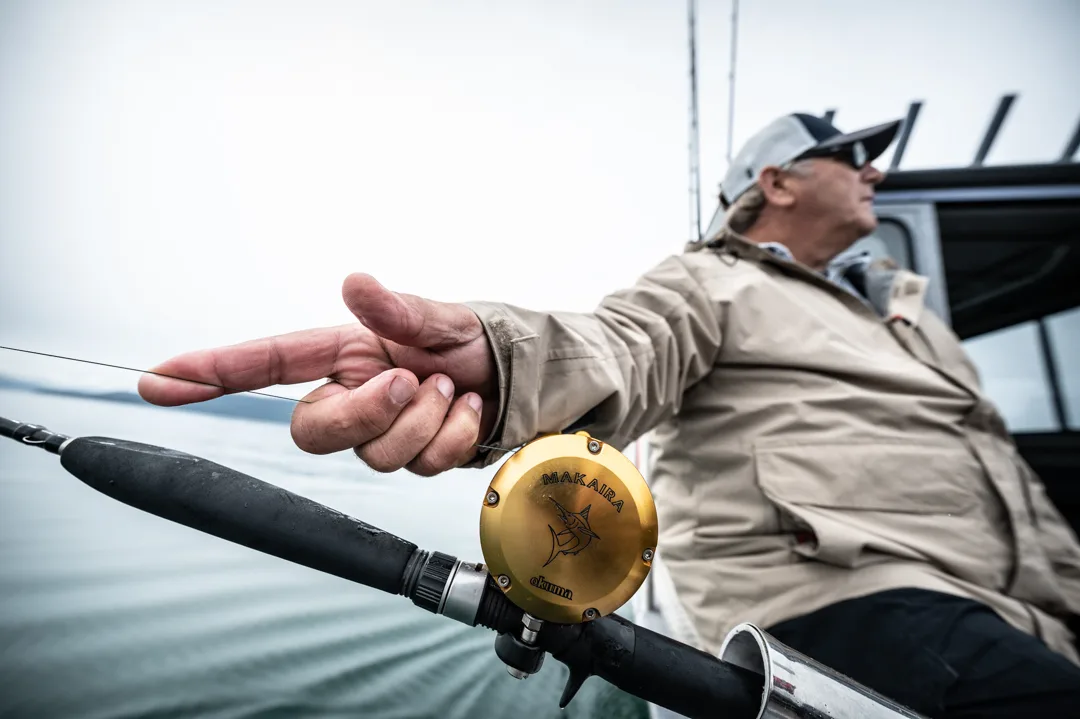 Angler pointing from the stern, Okuma Makaira gold reel and heavy rod in the foreground, calm water and mountains behind.