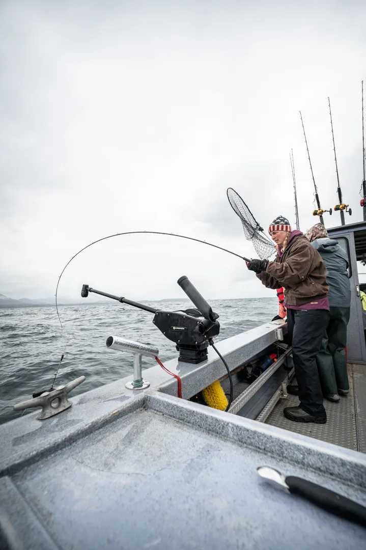 Angler in winter hat fights a fish on a bent rod, net ready, on the deck of a charter boat in overcast Icy Strait waters.