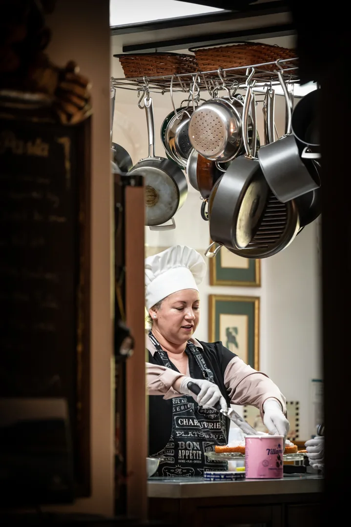 Chef in white toque and apron working at kitchen prep station, framed by hanging pots overhead