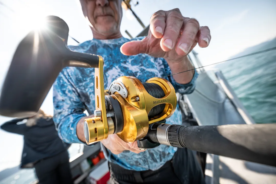 Angler checks line on a gold Penn Makara lever-drag reel aboard an open boat, calm water visible behind.