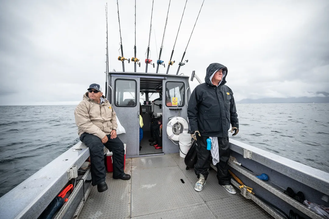 Two anglers on the deck of a charter boat, rain gear on, rods racked, open water and overcast sky behind them.