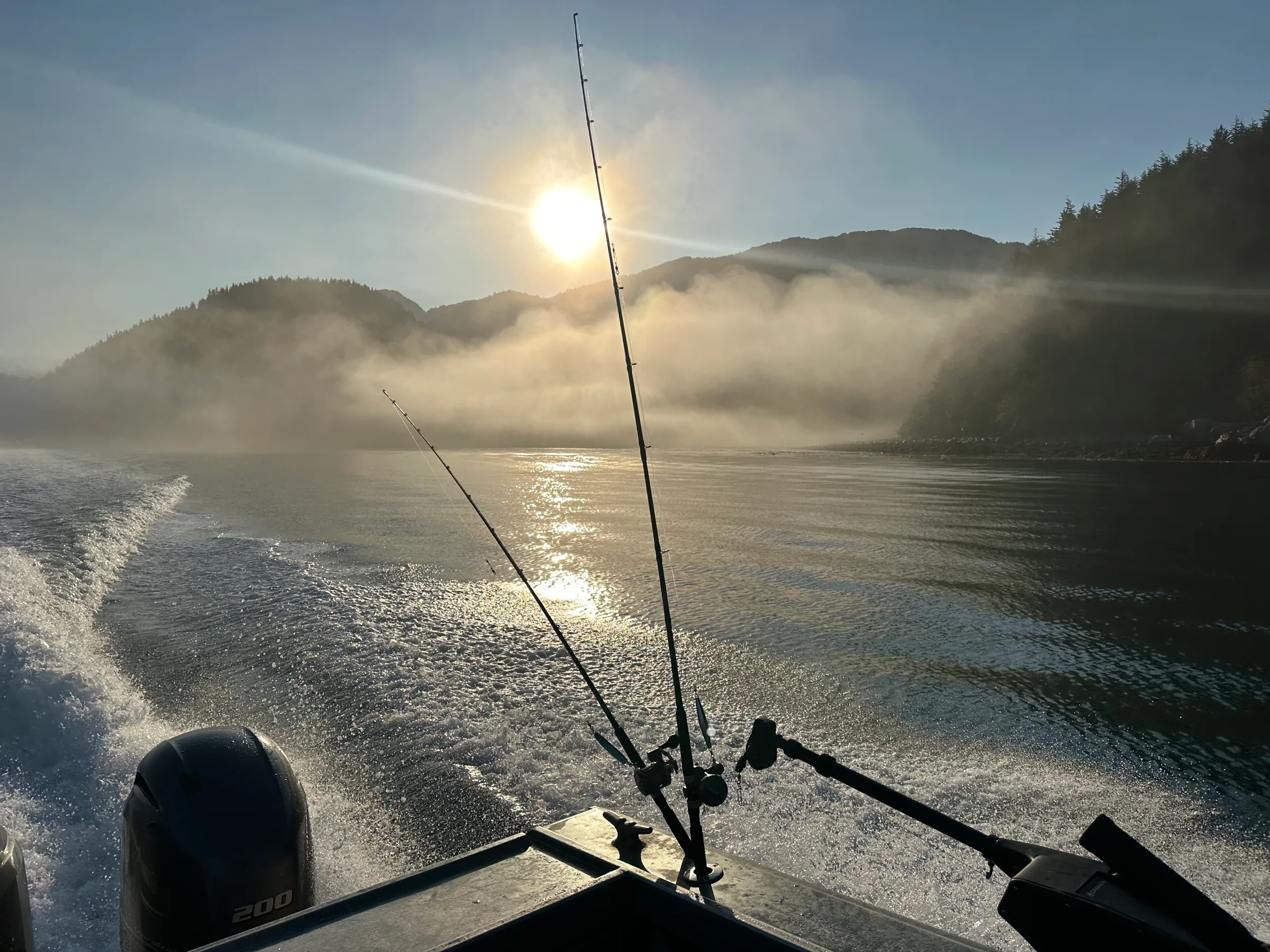 Fishing rods silhouetted against morning sun and low mist over mountain-flanked inlet, wake trailing behind outbound boat.