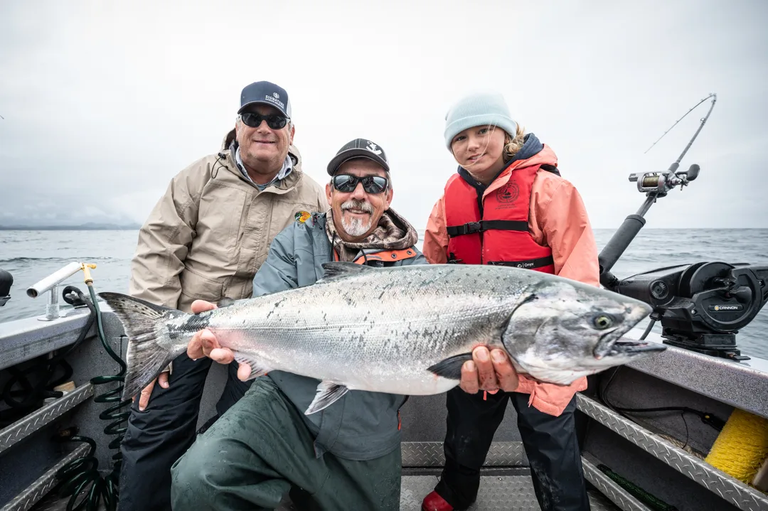 Two adults and a child on a fishing boat holding a large silver salmon, overcast skies and open water behind them.