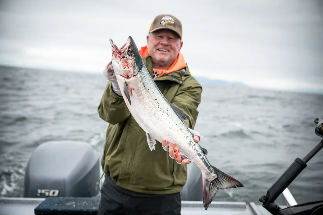 Angler holding a silver salmon on the back deck of a skiff, open water and overcast sky behind him.