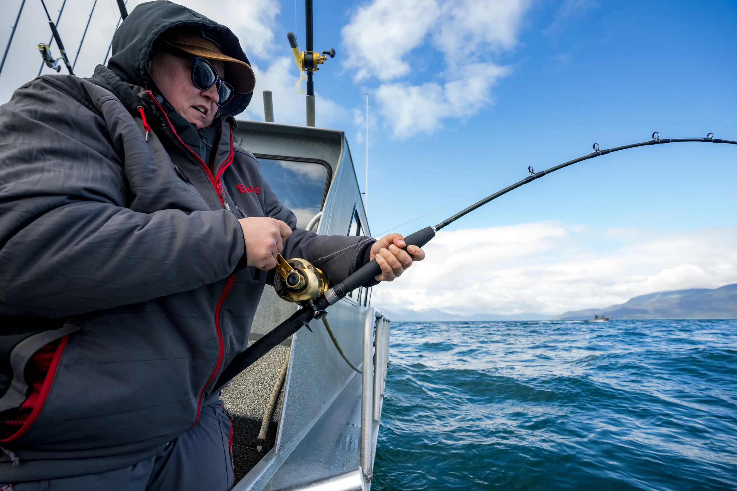 Angler in hooded jacket works a bent rod over open water, mountains visible on the horizon under a partly cloudy sky.