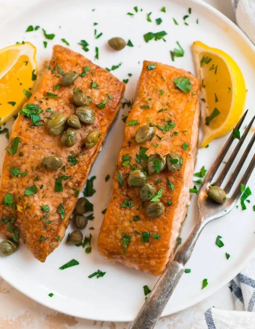 Two pan-seared salmon fillets topped with capers and chopped parsley, plated with lemon wedges on a white plate.