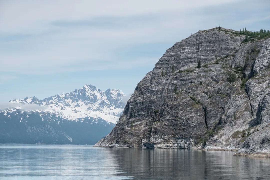 Steep granite cliff rises from calm water in Glacier Bay, snow-capped peaks visible in the distance.