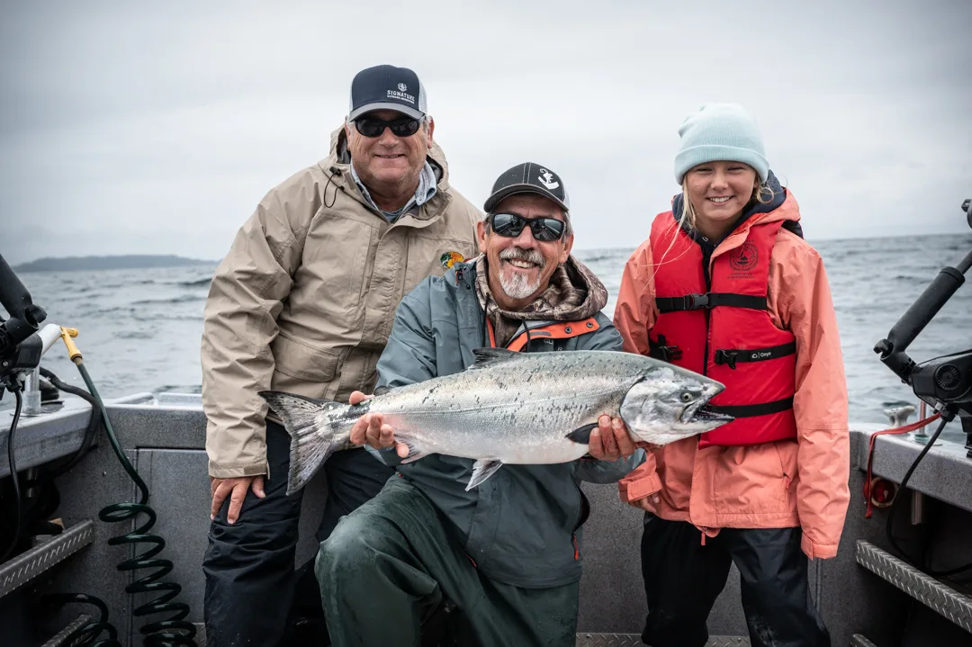 Three anglers on an aluminum boat holding a large silver salmon, overcast sky and open water behind them.