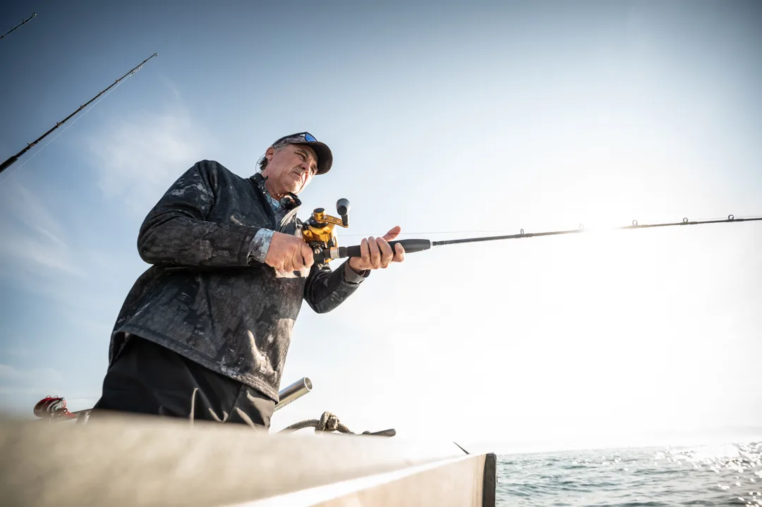 Angler braced at the gunwale, working a heavy rod with a gold conventional reel against an open sky.