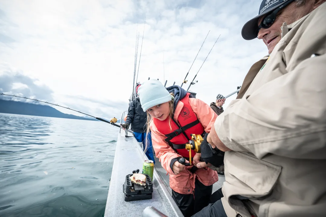 Young girl in red life vest works a gold reel alongside an adult angler on the deck of a charter boat, Icy Strait.