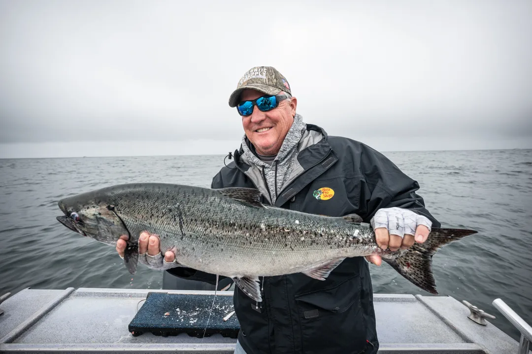 Angler holding a large king salmon on the boat deck, overcast sky and open water behind him.