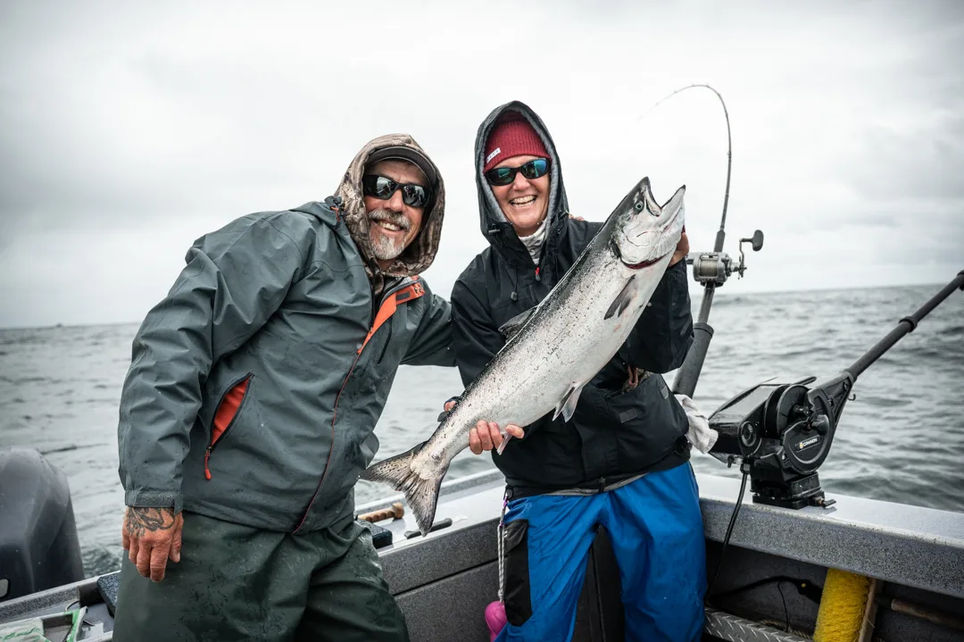 Two anglers in rain gear holding a large king salmon on a boat deck, overcast skies and open water behind them.
