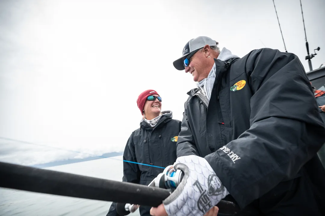 Two anglers in rain gear laughing on deck, rod in hand, mountains and gray water visible behind them.