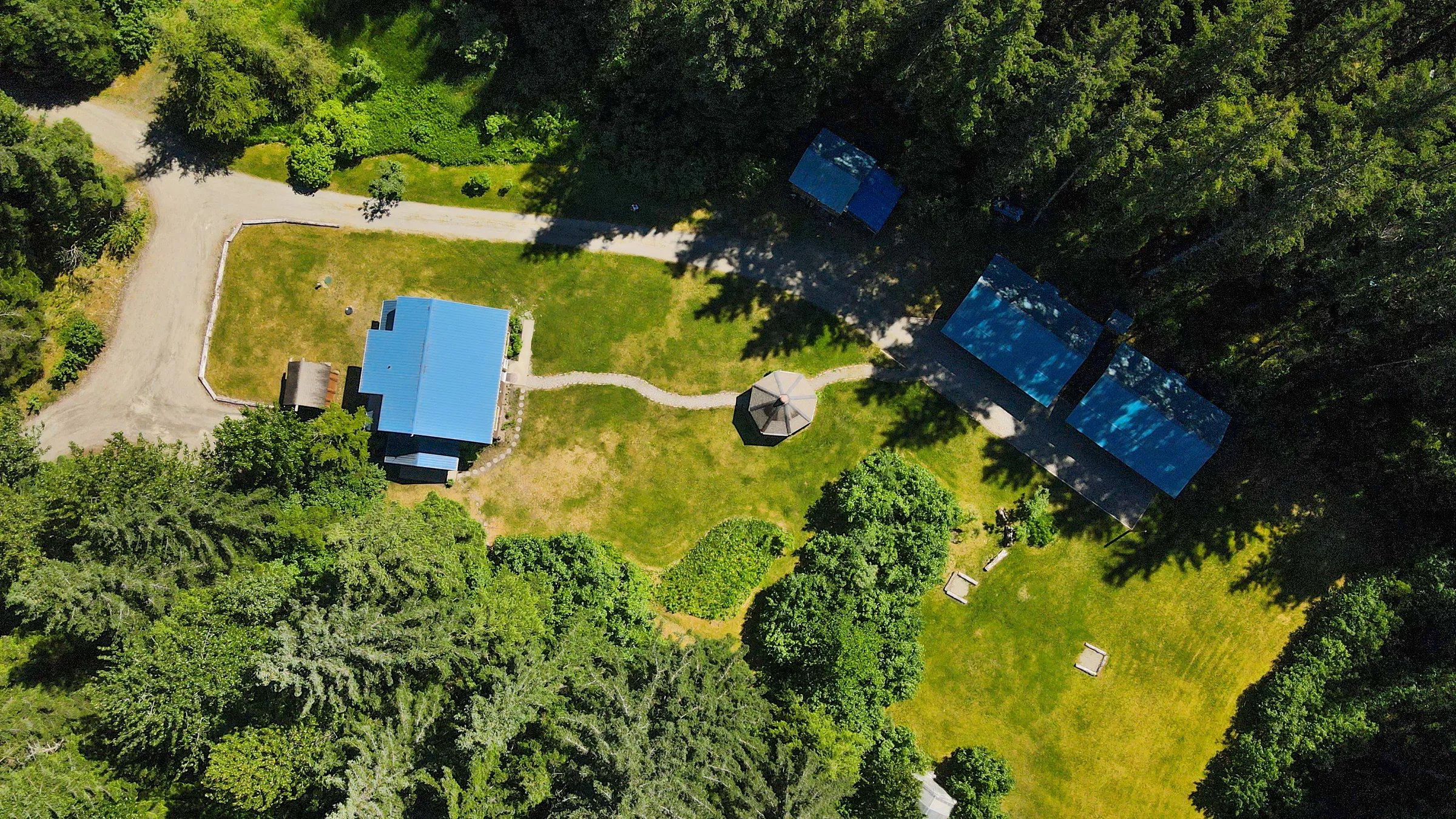 Aerial view of lodge buildings with blue metal roofs set among spruce forest and open lawn, connected by gravel paths.