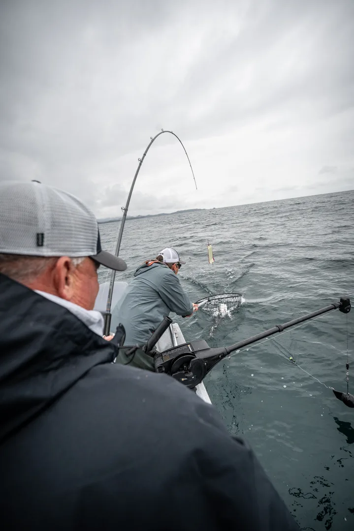 Angler nets a fish at the gunwale while a second rod bends under load, overcast Icy Strait in the background.
