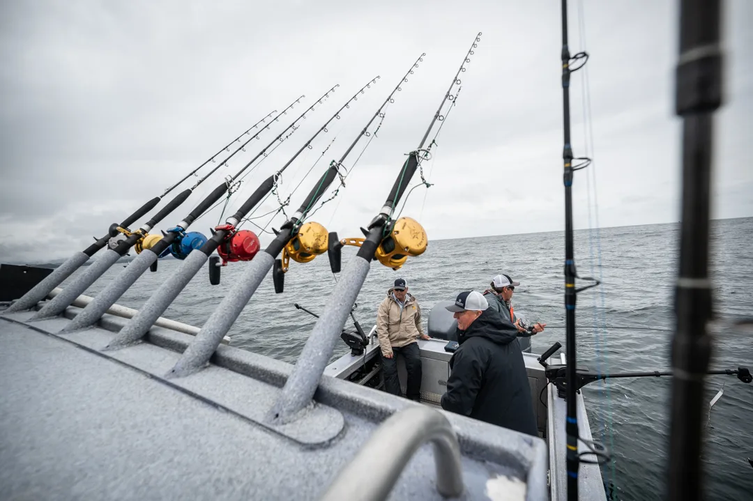 Row of heavy deep-sea rods racked on boat gunwale, three anglers on deck under overcast sky, open water behind.