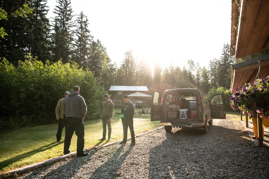 Guests and staff gathered on the gravel drive beside a van at the lodge, low sun cutting through spruce trees behind a cabin.