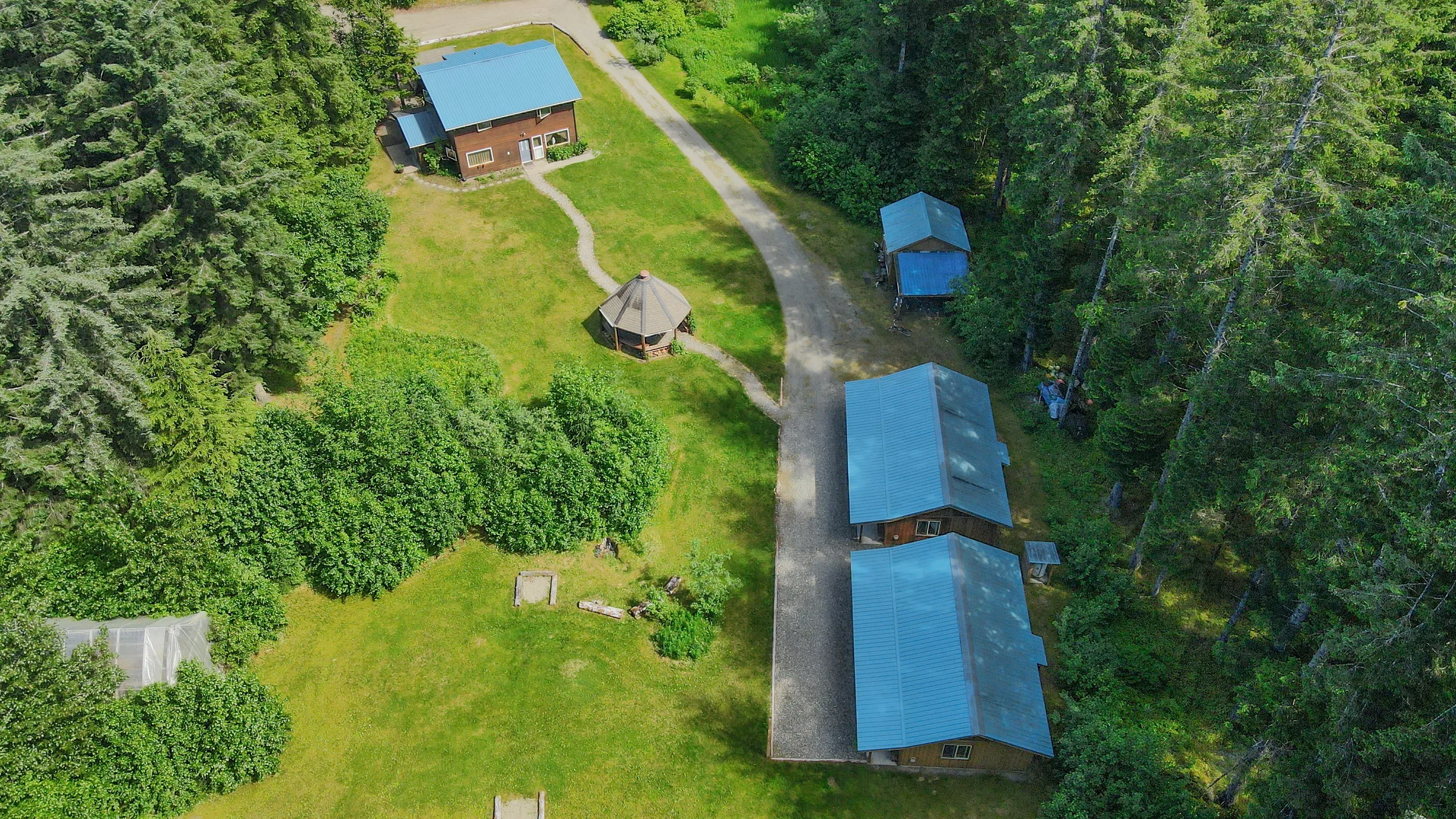 Aerial view of lodge compound — main building, guest cabins with blue metal roofs, and gazebo set among Sitka spruce forest.