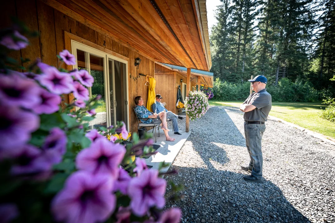 Two guests seated on cabin porch talking with a staff member, purple petunias in foreground, spruce forest behind.
