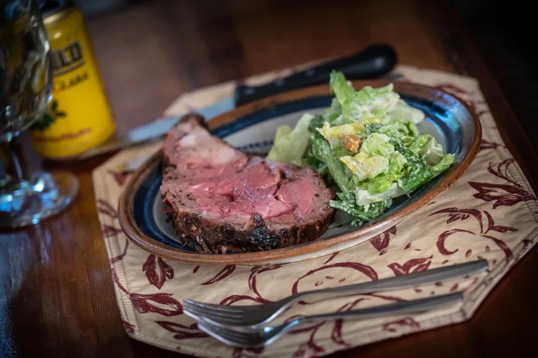 Sliced prime rib with Caesar salad on a ceramic plate, set on a patterned cloth placemat at a wood table.