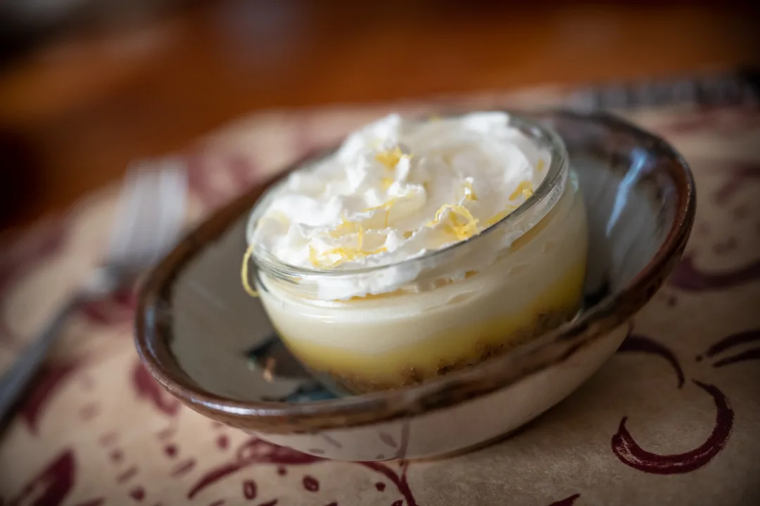 Lemon curd dessert in a glass jar topped with whipped cream and lemon zest, served in a handmade ceramic bowl.