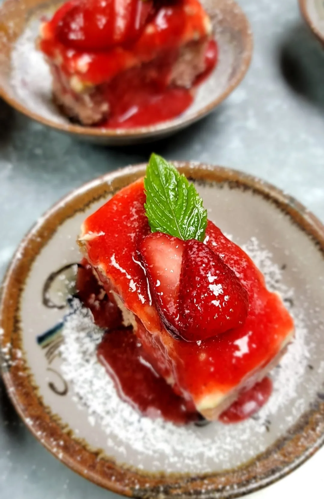Strawberry cheesecake bar with glazed berries and fresh mint on a rustic ceramic plate dusted with powdered sugar.