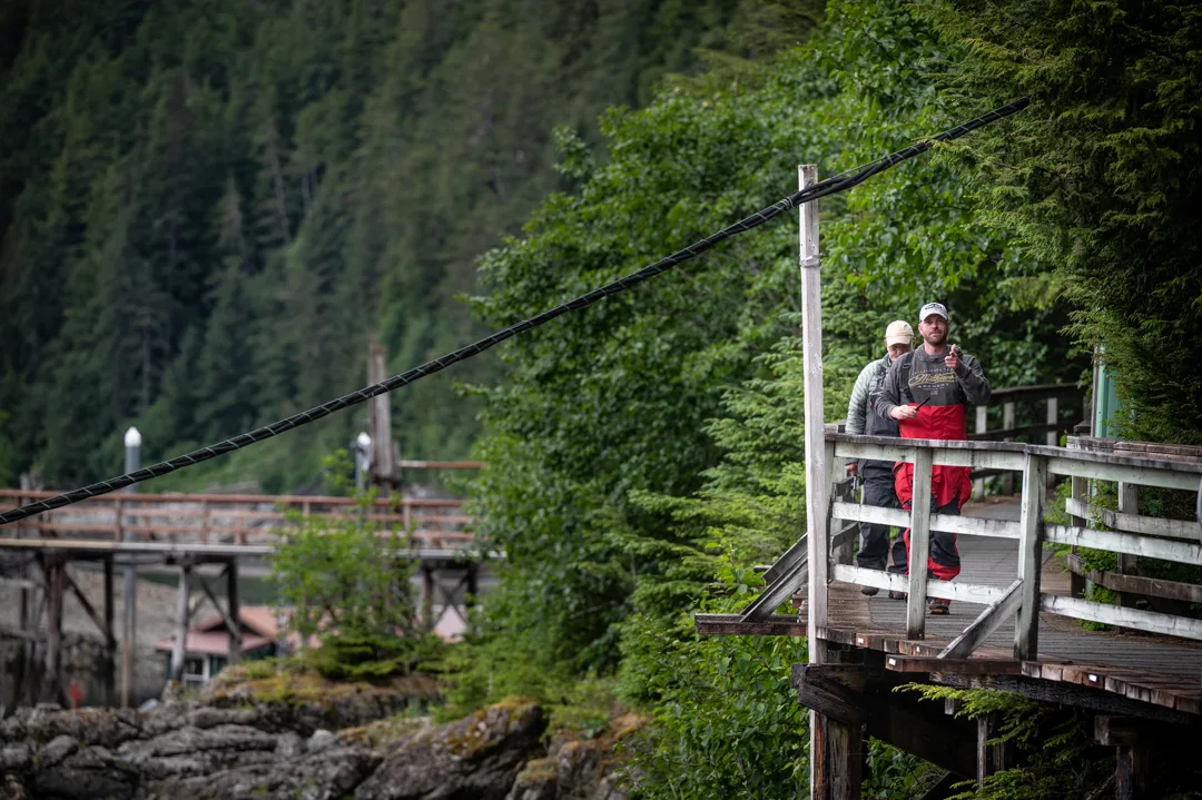 Two anglers in rain gear stand on a weathered wooden dock walkway, dense spruce forest and lodge structures visible behind.