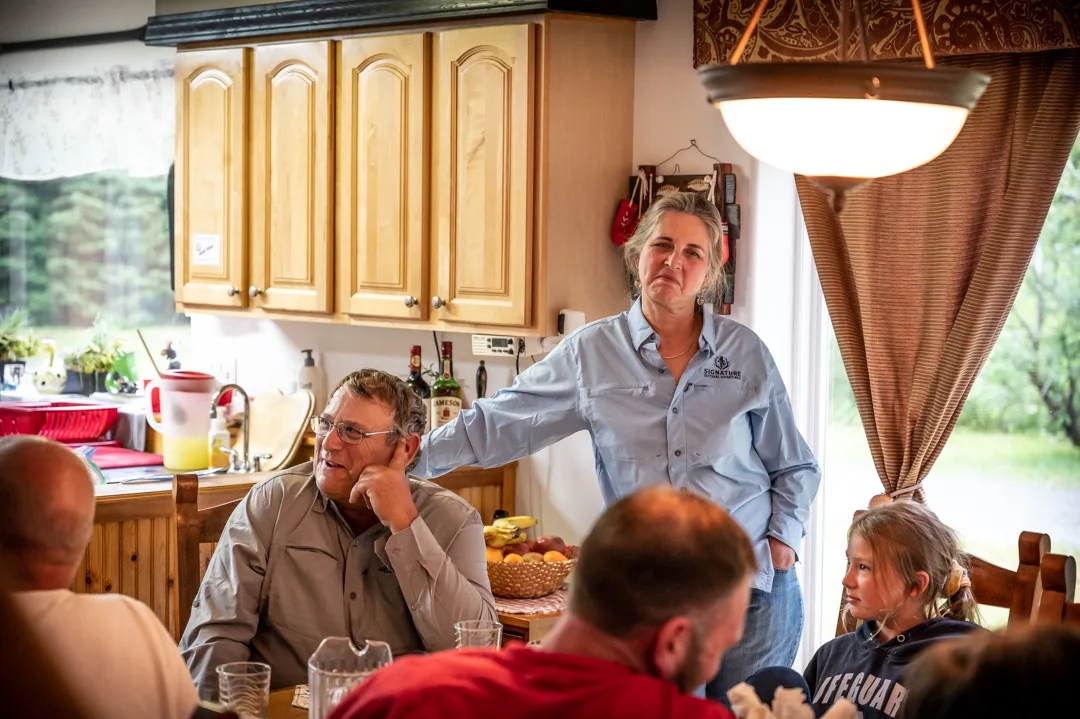 Lodge host speaks to guests gathered around a dining table inside the main lodge kitchen.