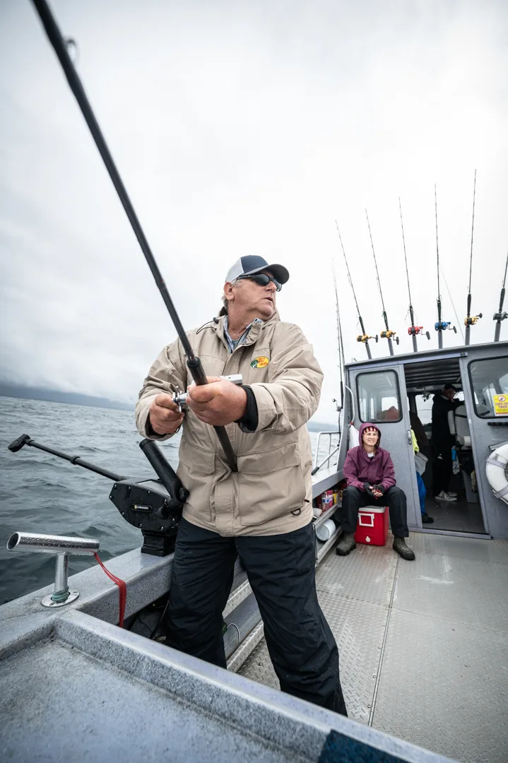 Angler working a bent rod on the stern deck of a charter boat, rod rack and overcast Icy Strait water in the background.