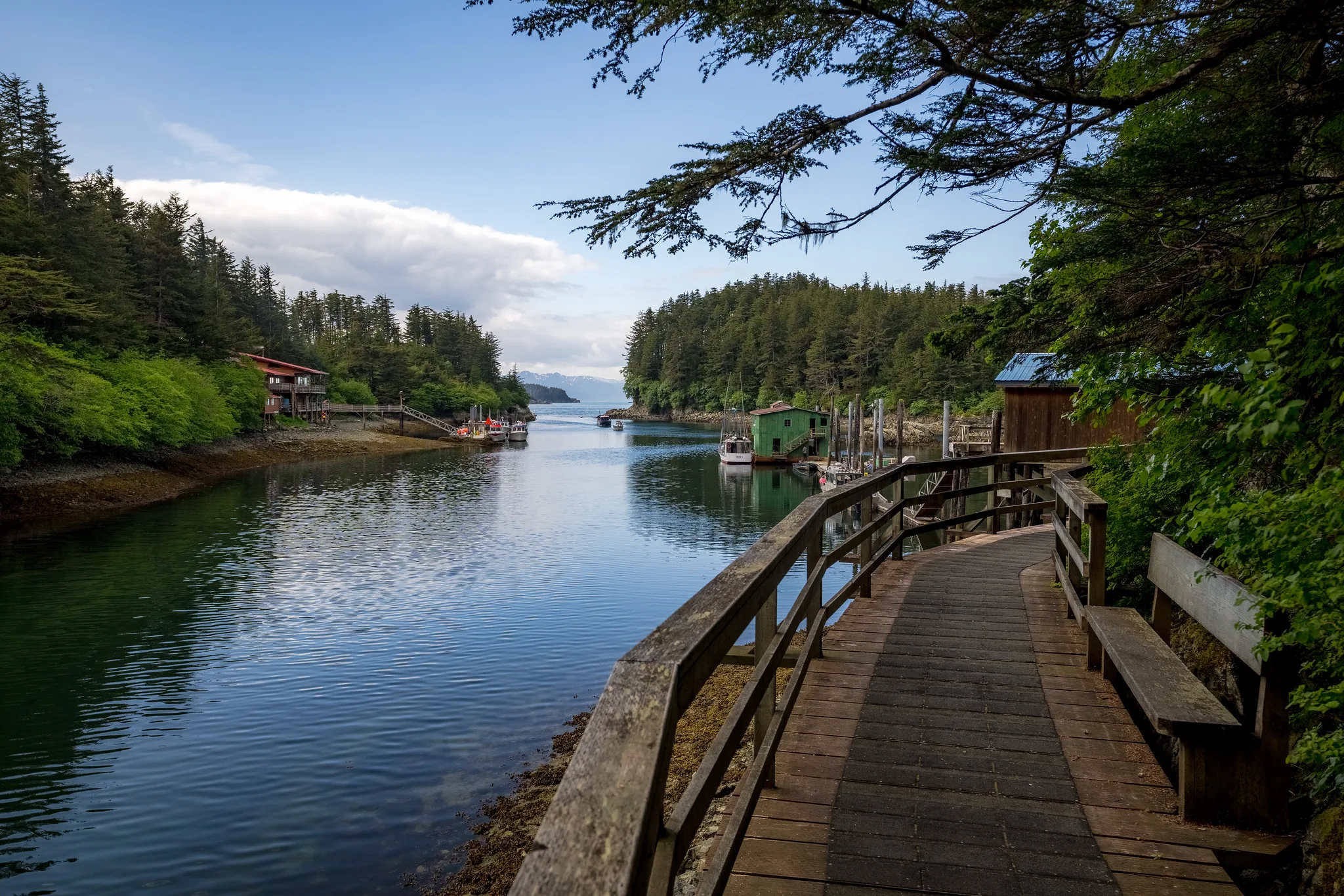 Wooden boardwalk curves along a calm inlet with docked boats, forested shoreline, and distant mountains under a partly cloudy sky.